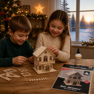 Bastelset „Wohnhaus“ aus naturbelassenem Holz mit einer Höhe von 12,5 cm. Auf einem Holztisch setzen zwei Kinder die einzelnen Holzteile eines detailreichen Wohnhauses zusammen. Neben dem teilweise aufgebauten Holzhaus liegen weitere Bauteile, Holzleim sowie die Verpackung des Bastelsets mit Hausabbildung. Im Hintergrund sorgen ein geschmückter Weihnachtsbaum, ein beleuchteter Stern und eine winterliche Landschaft am Fenster für eine gemütliche, festliche Atmosphäre. 