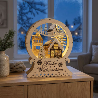LED-Diorama „Frohe Weihnachten“ aus naturfarbenem Holz, ca. 25 cm groß, mit detailreicher Winterszene aus Häusern, Tannenbäumen und Figuren, warmweiß beleuchtet und mit Schriftzug „Frohe Weihnachten“ im Sockel.