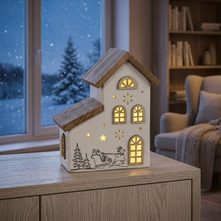 LED Keramikhaus mit Holzelementen, ca. 23 cm groß, mit beleuchteten Fenstern und ausgeschnittenen Sternmotiven, dekoriert mit weihnachtlicher Szene (Schlitten und Tannen), warmweiß beleuchtet, batteriebetrieben und mit integrierter Timerfunktion, auf einem Möbelstück vor winterlicher Fensterkulisse platziert.