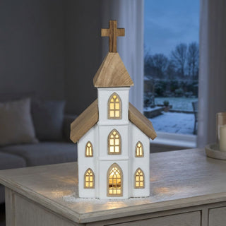LED Keramikkirche mit Holzelementen, ca. 23 cm groß, mit beleuchteten Fenstern und Kirchturm mit Holzkreuz, warmweiß beleuchtet, batteriebetrieben mit integrierter Timerfunktion, dekorativ auf einem Tisch vor winterlicher Fensterkulisse platziert.