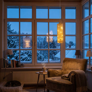 LED Glaszylinder zum Hängen in Amber-Gold, ca. 18 cm groß, mit integrierter Lichterkette, die ein warmweißes Licht abstrahlt, dekorativ von der Decke hängend vor großer Fensterfront mit winterlicher Schneelandschaft, in gemütlicher Wohnatmosphäre inszeniert.