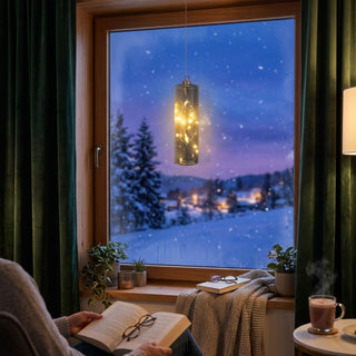 LED Glaszylinder zum Hängen in Grau, ca. 18 cm groß, mit integrierter warmweiß leuchtender Lichterkette, dekorativ vor einem Fenster mit verschneiter Winterlandschaft aufgehängt, umgeben von gemütlicher Wohnatmosphäre mit Vorhängen, Pflanzen und stillvoll arrangierter Dekoration.
