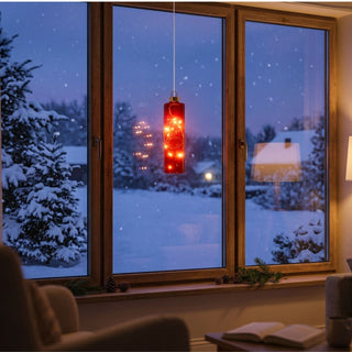 LED Glaszylinder zum Hängen in Rot, ca. 14 cm groß, mit integrierter warmweiß leuchtender Lichterkette, dekorativ vor einem Fenster mit verschneiter Winterlandschaft aufgehängt, in gemütlicher Wohnatmosphäre mit weihnachtlichen Dekorationselementen inszeniert.