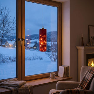 LED Glaszylinder zum Hängen in Rot, ca. 18 cm groß, mit integrierter warmweiß leuchtender Lichterkette, dekorativ vor einem Fenster mit verschneiter Winterlandschaft aufgehängt, in gemütlicher Wohnatmosphäre mit Sessel, Decke und Kamin im Hintergrund inszeniert.