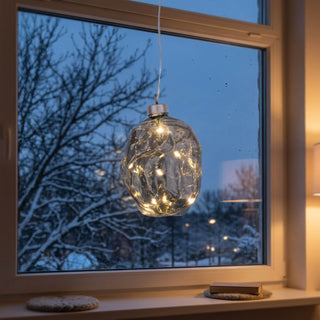 ED Glasnugget zum Hängen in Grau, ca. 10 cm groß, mit strukturierter Glasoberfläche und integrierter warmweiß leuchtender Lichterkette, dekorativ vor einem Fenster mit verschneiter Winterlandschaft aufgehängt, stimmungsvoll in einer gemütlichen Wohnumgebung inszeniert.