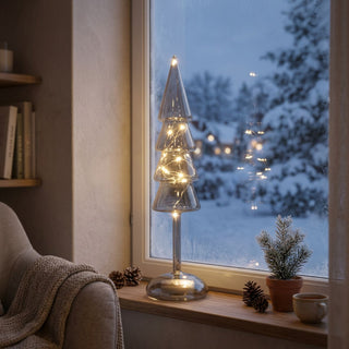 LED Dekoration „Weihnachtsbaum“ in Grau, ca. 32 cm groß, aus Glas gefertigt, in stilisierter Tannenbaumform mit mehreren übereinanderliegenden Ebenen, innen mit warmweiß leuchtender Lichterkette beleuchtet, dekorativ auf einer Fensterbank vor verschneiter Winterlandschaft platziert, umgeben von Tannenzapfen, kleiner Pflanze und gemütlichem Wohnambiente.