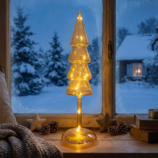 LED Glasdekoration „Weihnachtsbaum“ in Amber-Gold, ca. 32 cm groß, aus Glas gefertigt in stilisierter Tannenbaumform mit gestaffelten Ebenen, innen mit warmweiß leuchtender Lichterkette, auf einer Fensterbank vor verschneiter Winterlandschaft dekorativ platziert, umgeben von Tannenzapfen, Stern-Dekoelementen und gemütlicher Wohnatmosphäre.