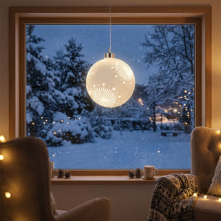 LED Glaskugel zum Hängen in Satin-Weiß, ca. 13 cm groß, mit dekorativem Stern- und Strahlenmuster, das durch die integrierte warmweiß leuchtende Beleuchtung hervorgehoben wird, vor einem Fenster mit verschneiter Winterlandschaft aufgehängt und in einer gemütlichen Wohnatmosphäre mit Sesseln, Decken und weihnachtlicher Dekoration inszeniert.