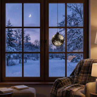 LED Glaskugel zum Hängen in Satin-Schwarz, ca. 13 cm groß, mit dekorativem Stern- und Strahlenmuster, das durch die integrierte warmweiß leuchtende Beleuchtung hervorgehoben wird, vor einem Fenster mit verschneiter Winterlandschaft aufgehängt und in einer gemütlichen Wohnatmosphäre mit Sessel, Decke und stimmungsvoller Beleuchtung inszeniert.