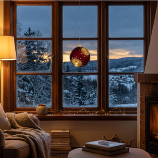 LED Glaskugel zum Hängen in Satin-Rot, ca. 13 cm groß, mit dekorativem Stern- und Strahlenmuster, das durch die integrierte warmweiß leuchtende Beleuchtung hervorgehoben wird, vor einem Fenster mit verschneiter Winterlandschaft aufgehängt und in einer gemütlichen Wohnatmosphäre mit Sessel, Decke, Bücherstapel und Kamin inszeniert.