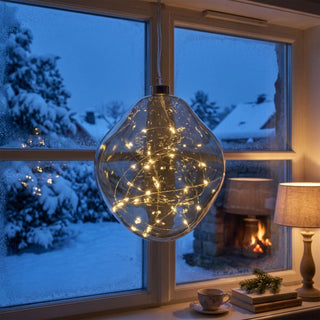 LED Glasform „organisch“ zum Hängen in Grau, ca. 19 cm groß, mit weich geschwungener, unregelmäßiger Form und integrierter warmweiß leuchtender Lichterkette, dekorativ vor einem Fenster mit verschneiter Winterlandschaft aufgehängt, stimmungsvoll in einer gemütlichen Wohnumgebung mit Lampe, Büchern, Tasse und Kaminfeuer im Hintergrund inszeniert.