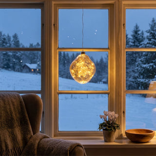 LED Glasform „organisch“ zum Hängen in Amber-Gold, ca. 19 cm groß, mit weich geschwungener, unregelmäßiger Form und integrierter warmweiß leuchtender Lichterkette, dekorativ vor einem Fenster mit verschneiter Winterlandschaft aufgehängt, stimmungsvoll in einer gemütlichen Wohnumgebung mit Sessel, Pflanze und warmem Licht inszeniert.