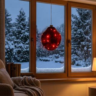LED Glasform „organisch“ zum Hängen in Rot, ca. 14 cm groß, mit weich geschwungener, unregelmäßiger Form und integrierter warmweiß leuchtender Lichterkette, dekorativ vor einem Fenster mit verschneiter Winterlandschaft aufgehängt, stimmungsvoll in einer gemütlichen Wohnumgebung mit Sessel, Decke und Büchern inszeniert.