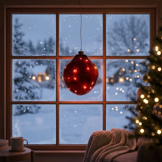 LED Glasform „organisch“ zum Hängen in Rot, ca. 19 cm groß, mit weich geschwungener, unregelmäßiger Form und integrierter warmweiß leuchtender Lichterkette, dekorativ vor einem Fenster mit verschneiter Winterlandschaft aufgehängt, stimmungsvoll in einer gemütlichen Wohnumgebung mit Sessel, Decke, Tasse und weihnachtlich beleuchtetem Tannenbaum im Hintergrund inszeniert.