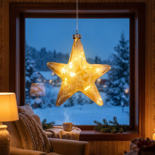 LED Glasstern zum Hängen in Amber-Gold, ca. 16 cm groß, in klassischer Sternform aus transparent getöntem Glas mit integrierter warmweiß leuchtender Lichterkette, dekorativ vor einem Fenster mit verschneiter Winterlandschaft aufgehängt, stimmungsvoll inszeniert in einer gemütlichen Wohnumgebung mit Sessel, Decke, Tasse und weihnachtlicher Dekoration.