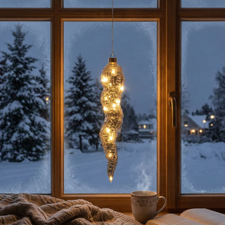 LED Glasstalaktit zum Hängen in Grau, ca. 27 cm groß, in länglicher, unregelmäßig strukturierter Form aus klarem bis leicht getöntem Glas, mit integrierter warmweiß leuchtender Lichterkette, die entlang der organischen Oberfläche schimmert, dekorativ vor einem Fenster mit verschneiter Winterlandschaft aufgehängt, stimmungsvoll inszeniert in einer gemütlichen Wohnumgebung mit Decke, Tasse und geöffnetem Buch auf der Fensterbank.