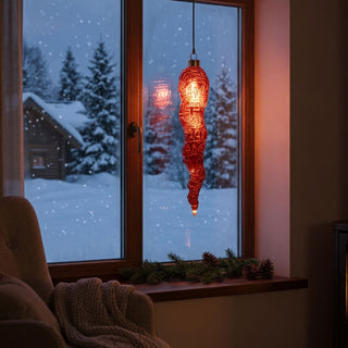 LED Glasstalaktit zum Hängen in Rot, ca. 27 cm groß, in länglicher, unregelmäßig strukturierter Form aus transparent rotem Glas, mit integrierter warmweiß leuchtender Lichterkette, die entlang der organischen Oberfläche schimmert, dekorativ vor einem Fenster mit verschneiter Winterlandschaft aufgehängt, stimmungsvoll inszeniert in einer gemütlichen Wohnumgebung mit Sessel, Decke, Tannenzweigen und sanfter Beleuchtung.