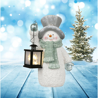XL Schneemann mit LED‑Laterne als Winterdekofigur, mit grauem Zylinder, grünem Schal und Handschuhen, hält eine warm leuchtende schwarze LED‑Laterne in der Hand, aufgestellt auf schneebedecktem Boden vor winterlichem Hintergrund mit Tannenbaum und blauem Bokeh‑Effekt, geeignet für stimmungsvolle Weihnachtsdekoration im Innen‑ und Außenbereich.