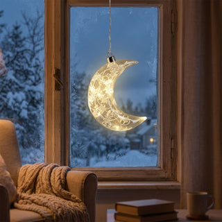 LED Glasmond mit Frost-Dekor, ca. 18 cm groß, in halbmondförmigem Design aus satiniertem Glas mit filigranem, frostähnlichem Muster, innen mit warmweiß leuchtender LED-Beleuchtung, dekorativ vor einem Fenster mit verschneiter Winterlandschaft aufgehängt, stimmungsvoll inszeniert in einer gemütlichen Wohnumgebung mit Sessel, Strickdecke, Büchern und sanfter Beleuchtung.