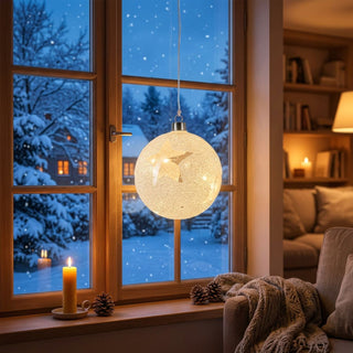 LED Glaskugel mit Frost-Dekor, ca. 12 cm Durchmesser, aus satiniertem Glas mit feiner, frostähnlicher Struktur, innen mit warmweiß leuchtender LED-Lichterkette, batteriebetrieben mit integrierter Timerfunktion, dekorativ an einem Fenster vor verschneiter Winterlandschaft aufgehängt, stimmungsvoll inszeniert in einer gemütlichen Wohnumgebung mit Kerzen, Tannenzapfen, Decke und Sofa.