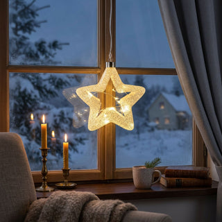 LED Glasstern zum Hängen mit Frost-Dekor, ca. 15 cm groß, in klassischer Sternform aus satiniertem Glas mit feiner, frostähnlicher Struktur, innen mit warmweiß leuchtender Lichterkette, dekorativ vor einem Fenster mit verschneiter Winterlandschaft aufgehängt, stimmungsvoll inszeniert in einer gemütlichen Wohnumgebung mit Vorhang, Kerzen, Tasse, Büchern und weihnachtlicher Dekoration auf der Fensterbank.