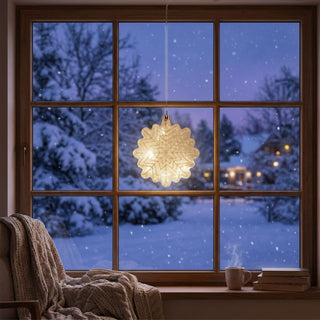 LED „Schneeflocke“ aus Glas (Ø 15 cm) in Kristalloptik, warmweiß beleuchtet, als elegante Fensterdekoration in gemütlicher Winteratmosphäre mit verschneiter Landschaft im Hintergrund.