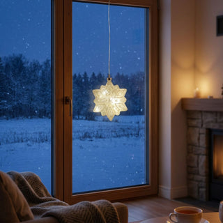 LED „Schneeflocke“ aus Glas (Ø 15 cm) in Kristalloptik, warmweiß beleuchtet, als hängende Fensterdekoration vor winterlicher Landschaft mit Kamin und gemütlichem Wohnambiente im Hintergrund.