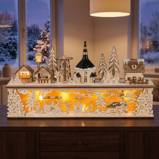 LED-Lichterhaus „Winterdorf“ (45 cm) mit Sockel, detailreiche Holzdekoration mit warm leuchtender Innenbeleuchtung. Auf der Oberseite sind kleine Häuser, Bäume und eine Kirche angeordnet, während im unteren Bereich eine filigrane Winterszene mit Figuren und verschneiter Landschaft dargestellt ist. Die Dekoration steht auf einem Möbelstück vor einem Fenster mit winterlichem Ausblick und sorgt für eine gemütliche, festliche Atmosphäre im Innenraum.