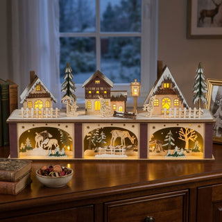 LED-Lichterhaus „Winterstadt“ (45 cm) auf Sockel, farbige Holzszene mit beleuchteten Häusern, Bäumen und winterlichen Tiermotiven, warm leuchtend auf einer Kommode vor Fenster platziert.