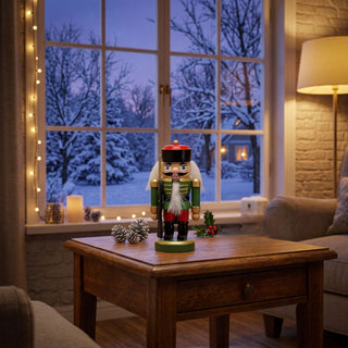 Nussknacker Mini, 10 cm groß, in grüner Lackierung, mit schwarzer Mütze, weißem Bart und Schnurrbart sowie goldfarbenen Details, dekorativ auf einem Tisch vor winterlicher Fensterkulisse mit Lichterkette, Kerzenlicht und gemütlicher Wohnatmosphäre platziert.