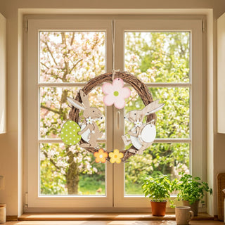 Fensterbild „Osterhase“ (ca. 22 cm), dekorativer Türkranz mit zwei Hasenfiguren, Blüten und bunten Eiern, aufgehängt am Fenster. Im Hintergrund ein blühender Garten, auf der Fensterbank stehen kleine Töpfe mit Kräutern.