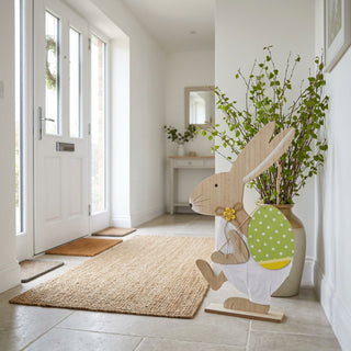 Osterhase XL (ca. 60 cm) in Natur-Optik, dekorativer Holzaufsteller mit angedeuteter Stoffhose und großem gepunkteten Osterei. Steht im Eingangsbereich neben einer Vase mit Zweigen, mit Blick zur Haustür und heller Wohnumgebung.