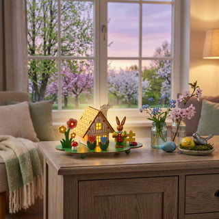 LED-Lichterhaus „Blumenwiese“ (ca. 9 cm), farbiges Minihaus mit leuchtenden Fenstern, umgeben von Blumen und kleinem Osterhasen. Platziert auf einer Kommode neben Vasen mit Frühlingsblumen und Ostereiern, vor einem Fenster mit Blick in einen blühenden Garten bei Dämmerung.