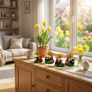 Osterhasen mit Blume (4er-Set, je ca. 6,5 cm), kleine farbig sortierte Hasenfiguren auf grünen Sockeln mit Blumen und Ostereiern. Arrangiert auf einer Kommode mit Tulpen, Narzissen und Osterdeko vor einem Fenster mit Blick in einen blühenden Frühlingsgarten.