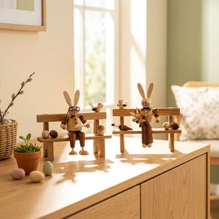 Osterhasenpaar auf Bank (ca. 13 cm), naturfarbene Holzfiguren mit zwei Hasen auf separaten kleinen Holzbänken, dekoriert mit Vögeln und kleinen Eiern. Platziert auf einer Kommode neben Pflanzen und Osterdeko vor einem hellen Fenster.