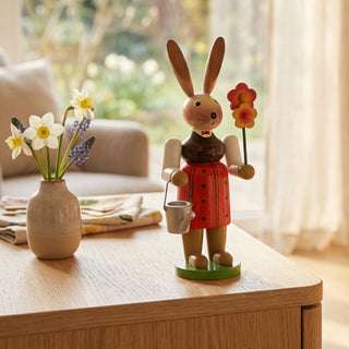 Osterhase „Gärtnerin“ (ca. 30 cm), farbig gestaltete Figur im roten Kleid, hält eine Blume und eine kleine Gießkanne. Platziert auf einer Holzkommode neben Vase mit Frühlingsblumen vor einem Fenster mit Blick in einen blühenden Garten.