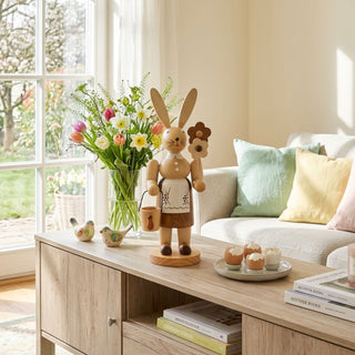 Osterhase „Gärtnerin“ (ca. 30 cm), naturfarbene Holzfigur mit Schürze, Blume und kleinem Eimer in der Hand. Steht auf einer Kommode neben Vase mit Frühlingsblumen und Kerzen, vor einem Fenster mit Blick in einen blühenden Garten.