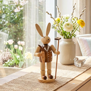 Osterhase „Gärtner“ (ca. 30 cm), naturfarbene Holzfigur mit Weste, hält eine Möhre und einen Rechen. Platziert auf einer Kommode neben Vase mit Frühlingsblumen, vor einem Fenster mit Blick in einen blühenden Garten.