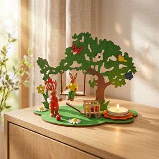 Kerzenhalter für Teelicht „Osterhase auf Schaukel“ (ca. 14,5 cm), farbiges Holz-Diorama mit Baum, schaukelndem Hasen, Blumen und kleinem Wagen, daneben ein brennendes Teelicht. Platziert auf einer Kommode in heller Wohnatmosphäre.