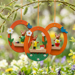 Osterhasen im Ei zum Hängen (3er-Set, je ca. 7 cm), drei farbige Anhänger in orangefarbenen Ei-Rahmen mit kleinen Hasenfiguren, Blumen und Vögeln. Hängen an Zweigen in einem blühenden Frühlingsgarten mit grünen Blättern und bunten Blumen im Hintergrund.