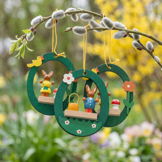 Osterhasen im Ei zum Hängen (3er-Set, je ca. 7 cm), drei grüne, eiförmige Anhänger mit kleinen Hasenfiguren, Blumen und Pilzdetails. Hängen an Zweigen in einem blühenden Frühlingsgarten mit weichem Hintergrund aus Grün- und Gelbtönen.