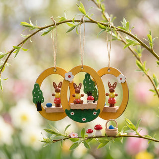 Osterhasen im Ei zum Hängen (3er-Set, je ca. 7 cm), drei gelbe, eiförmige Anhänger mit kleinen Hasenfiguren, Blumen- und Pilzmotiven. Hängen an Zweigen in einem blühenden Frühlingsgarten mit zarten Grün- und Pastellfarben im Hintergrund.