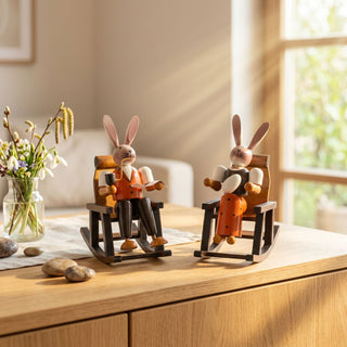 Osterhasenpaar „Oma und Opa im Schaukelstuhl“ (ca. 15 cm), zwei farbig gestaltete Hasenfiguren sitzend in Schaukelstühlen. Platziert auf einer Kommode neben Vase mit Frühlingsblumen in heller Wohnatmosphäre.