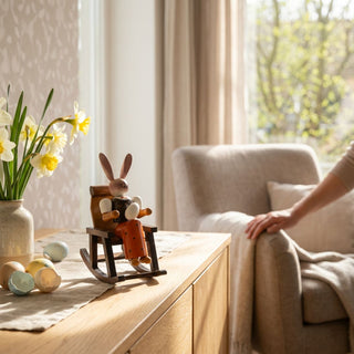 Osterhase „Oma im Schaukelstuhl“ (ca. 15 cm), farbig gestaltete Hasenfigur in einem Schaukelstuhl. Platziert auf einer Holzkommode neben Vase mit Narzissen und Ostereiern in heller Wohnatmosphäre.