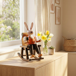 Osterhase „Opa im Schaukelstuhl“ (ca. 15 cm), farbig gestaltete Hasenfigur mit Pfeife sitzend in einem Schaukelstuhl. Platziert auf einer Holzkommode neben Vase mit Frühlingsblumen in heller Wohnatmosphäre.