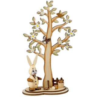 Dekorative Holzszene mit einem aufrecht stehenden Hasen in Weste und Knöpfen im Vordergrund. Dahinter ein großer Baum mit grünen Blättern und weißen Blüten. Auf einem Ast sitzt ein Vogel, in der Mitte des Baumes ein Schmetterling. Am Baumfuß liegen bunte Eier und ein kleiner Holzeimer.