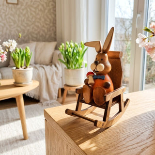 Osterhase im Schaukelstuhl (ca. 15 cm), farbig gestaltete Figur mit orangem Oberteil und Fliege, sitzend in einem Holz-Schaukelstuhl und ein kleines Objekt in der Hand haltend. Platziert auf einer Holzkommode neben blühenden Pflanzen in heller Wohnatmosphäre.