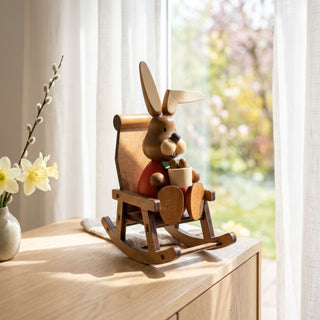 Osterhase im Schaukelstuhl (ca. 15 cm), farbig gestaltete Figur mit rotem Oberteil, sitzend in einem Holz-Schaukelstuhl und eine Tasse in den Händen haltend. Platziert auf einer Kommode neben Vase mit Narzissen vor einem Fenster mit Blick in einen blühenden Garten.