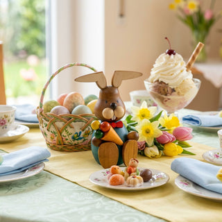 Osterhase mit Eis (ca. 13 cm), farbig gestaltete Figur mit blauem Oberteil, hält ein Eis in der Hand. Platziert auf einem gedeckten Tisch mit Osterdekoration, Blumen, bunten Eiern und Dessert im Hintergrund.