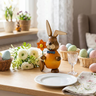 Osterhase mit Blume (ca. 13 cm), farbig gestaltete Figur mit orangem Oberteil, hält eine orange Blüte in der Hand. Platziert auf einem gedeckten Tisch mit Ostereiern, Körbchen und weißen Blumen in heller Wohnatmosphäre.